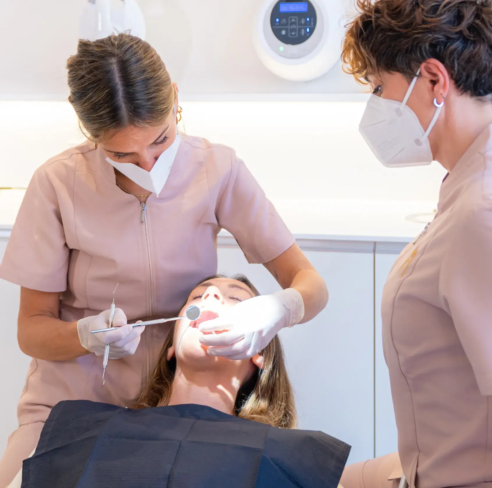 A dentist wearing a mask and gloves examines a patient's teeth with dental tools while an assistant observes.