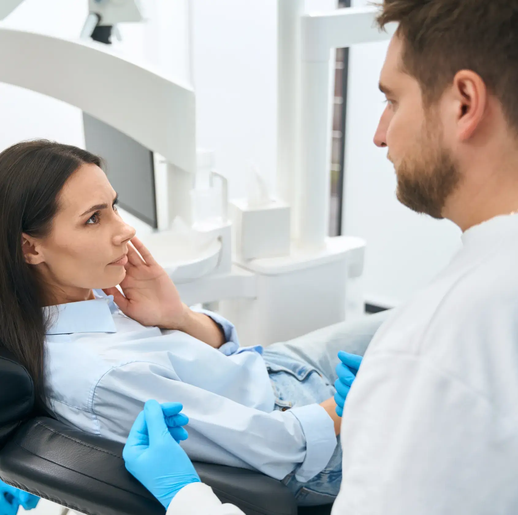 Woman sitting in dental chair looking concerned while a dentist wearing blue gloves examines her.