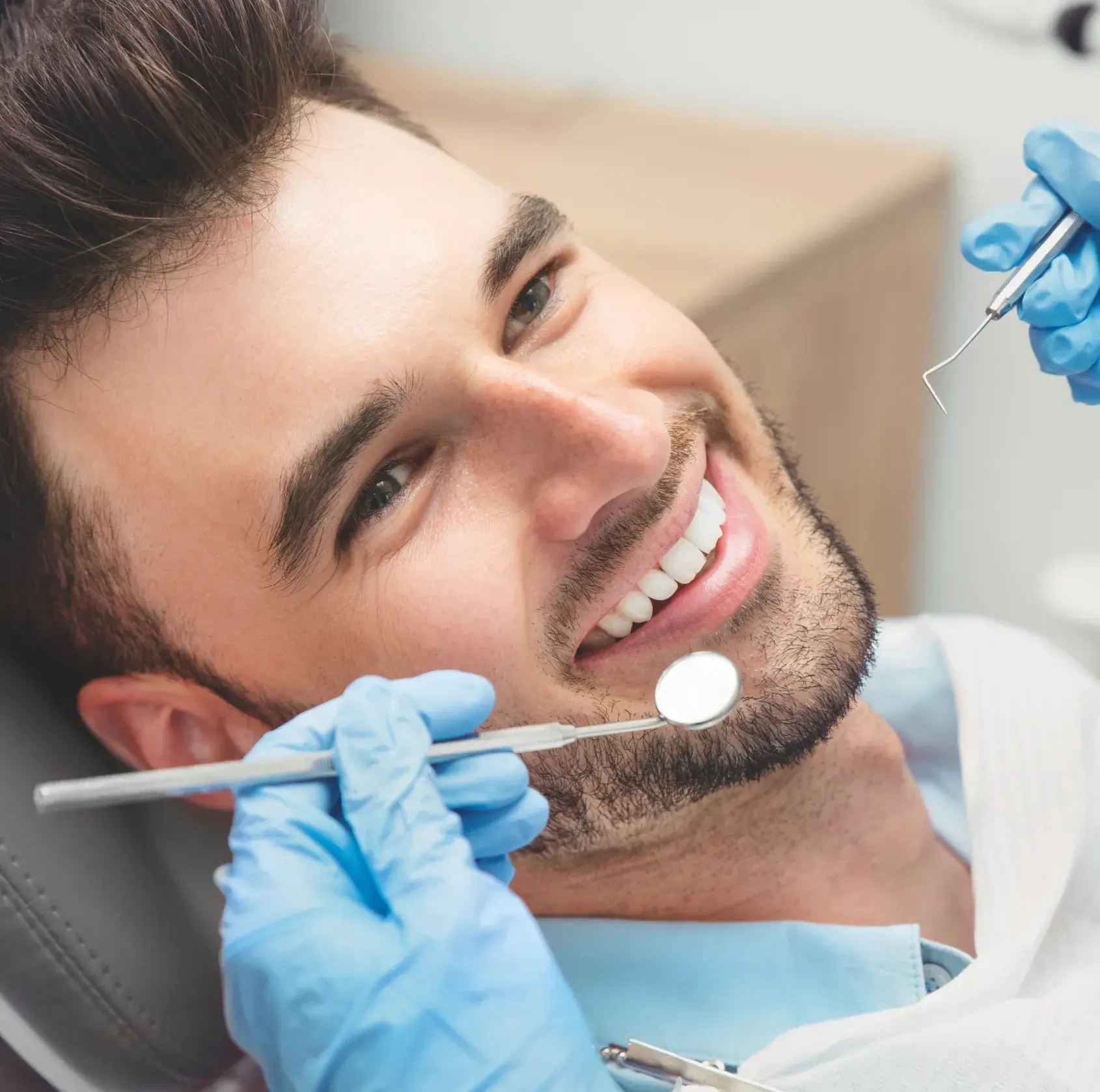 Smiling man reclined in dentist chair with dental mirror and probe held by gloved hands near his mouth.