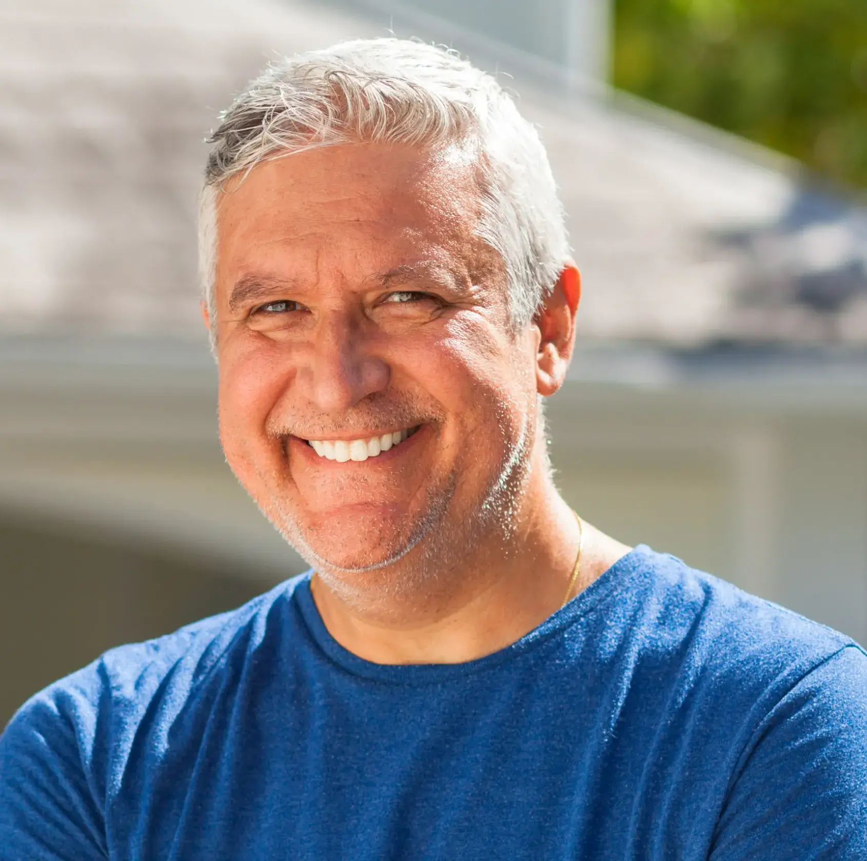 Middle-aged man with gray hair smiling brightly outdoors wearing a blue shirt.