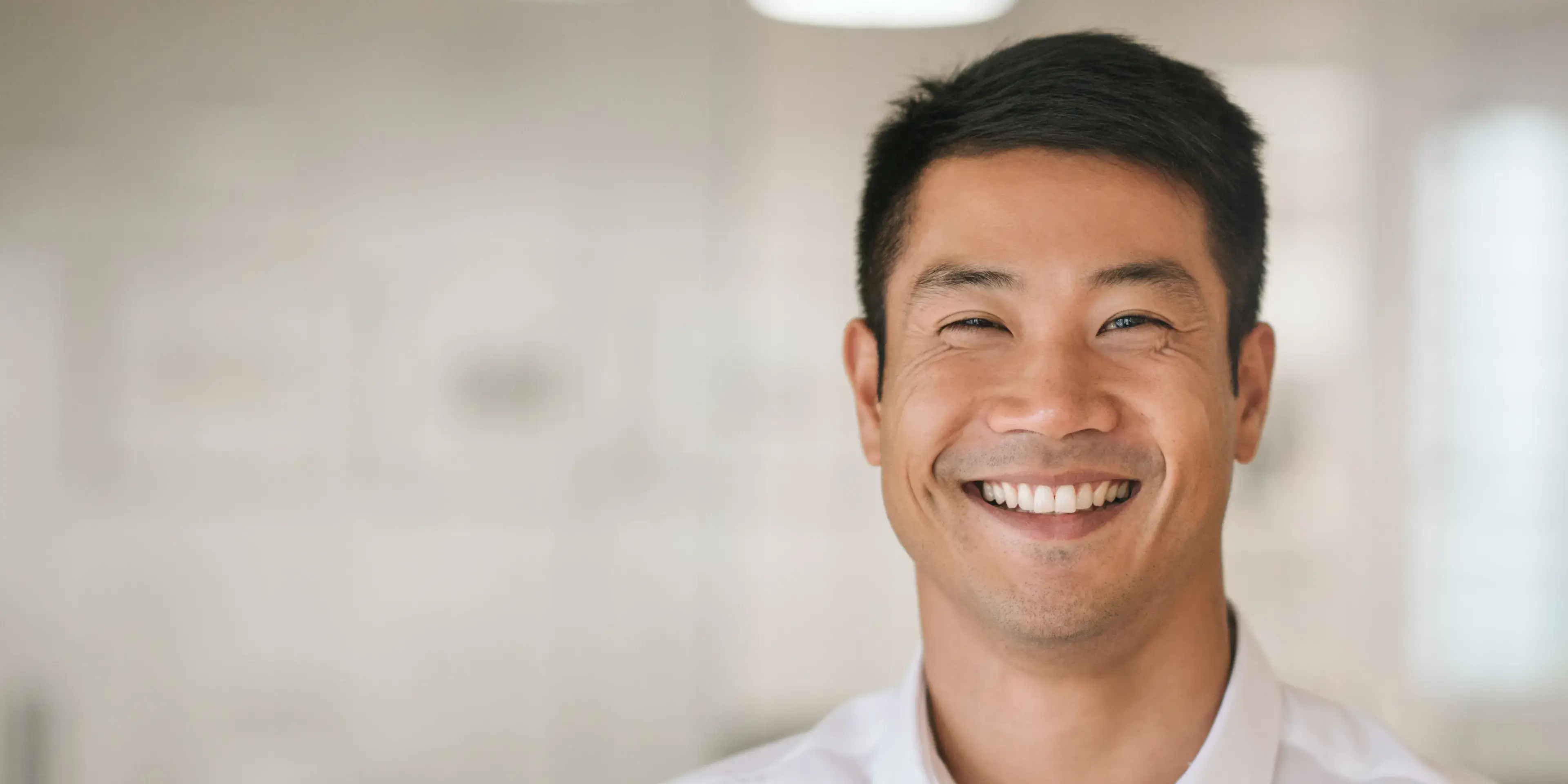 Smiling Asian man with short black hair showing bright white teeth in a softly lit indoor setting.