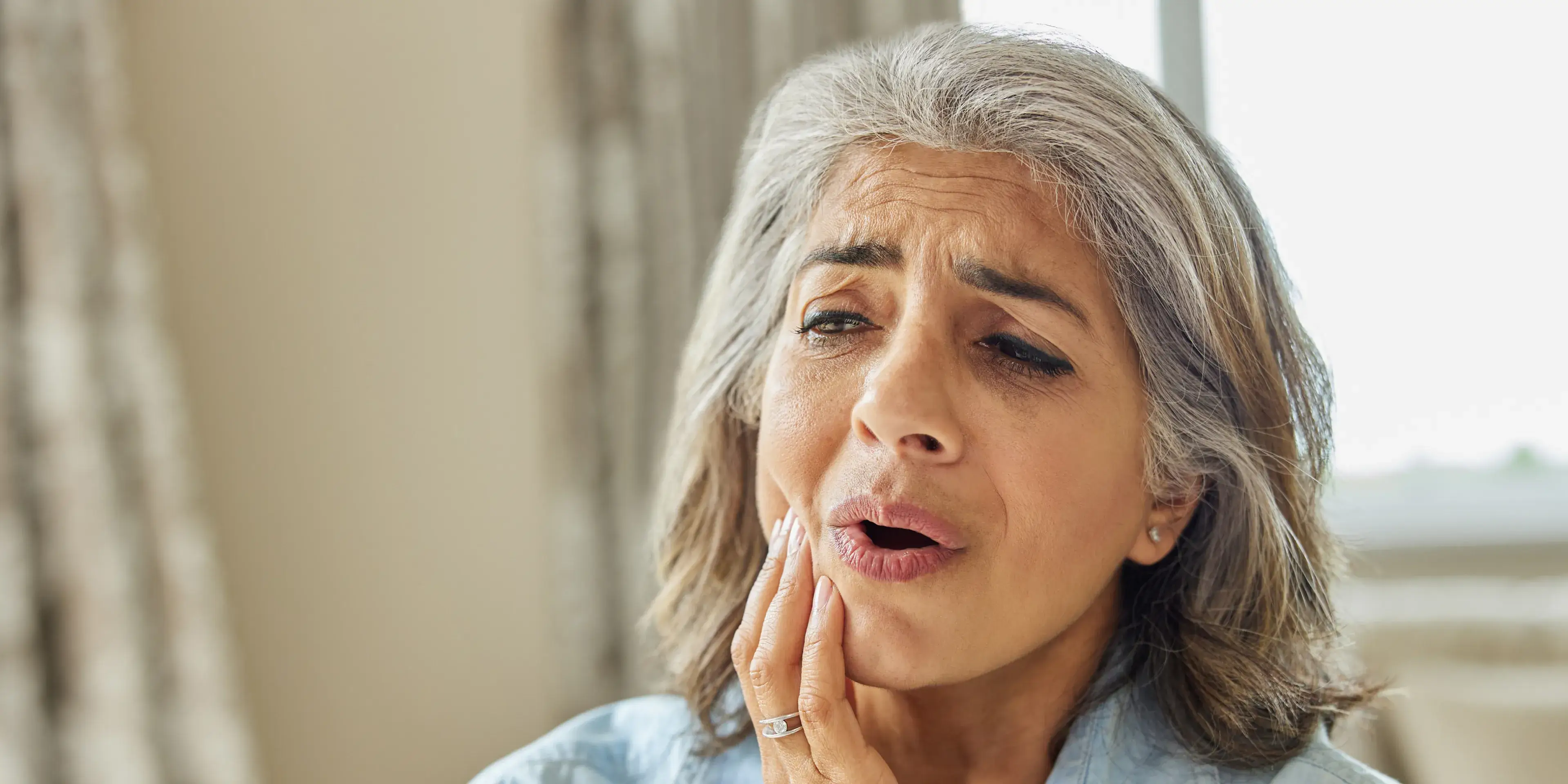 Middle-aged woman with gray hair holding her cheek, showing discomfort or tooth pain.