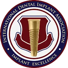Emblem of the International Dental Implant Association featuring a gold dental implant centered on a maroon shield with blue circular border containing the text 'International Dental Implant Association' and 'Implant Excellence'.