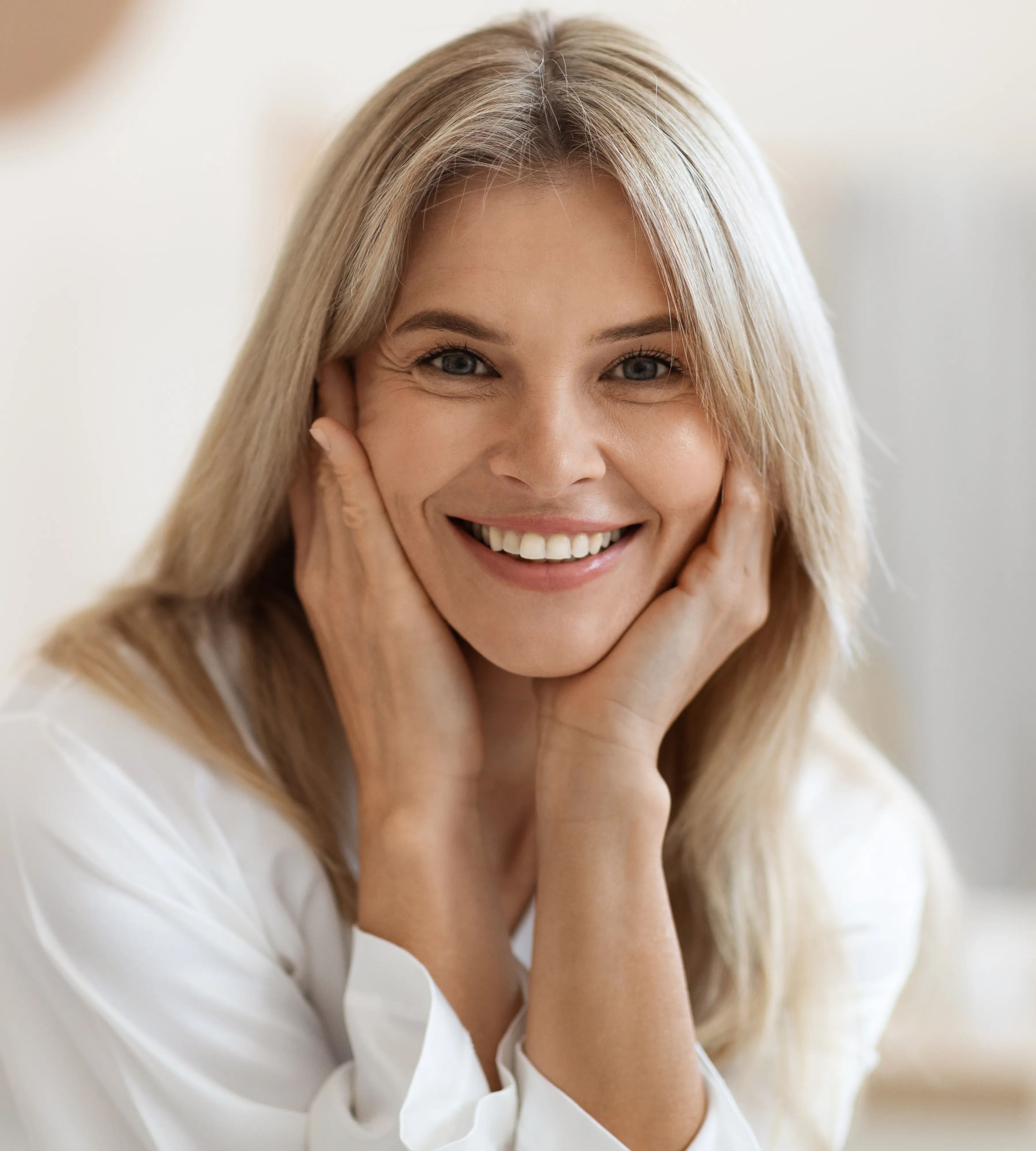 Smiling middle-aged woman with blonde hair resting her face in her hands, wearing a white shirt.