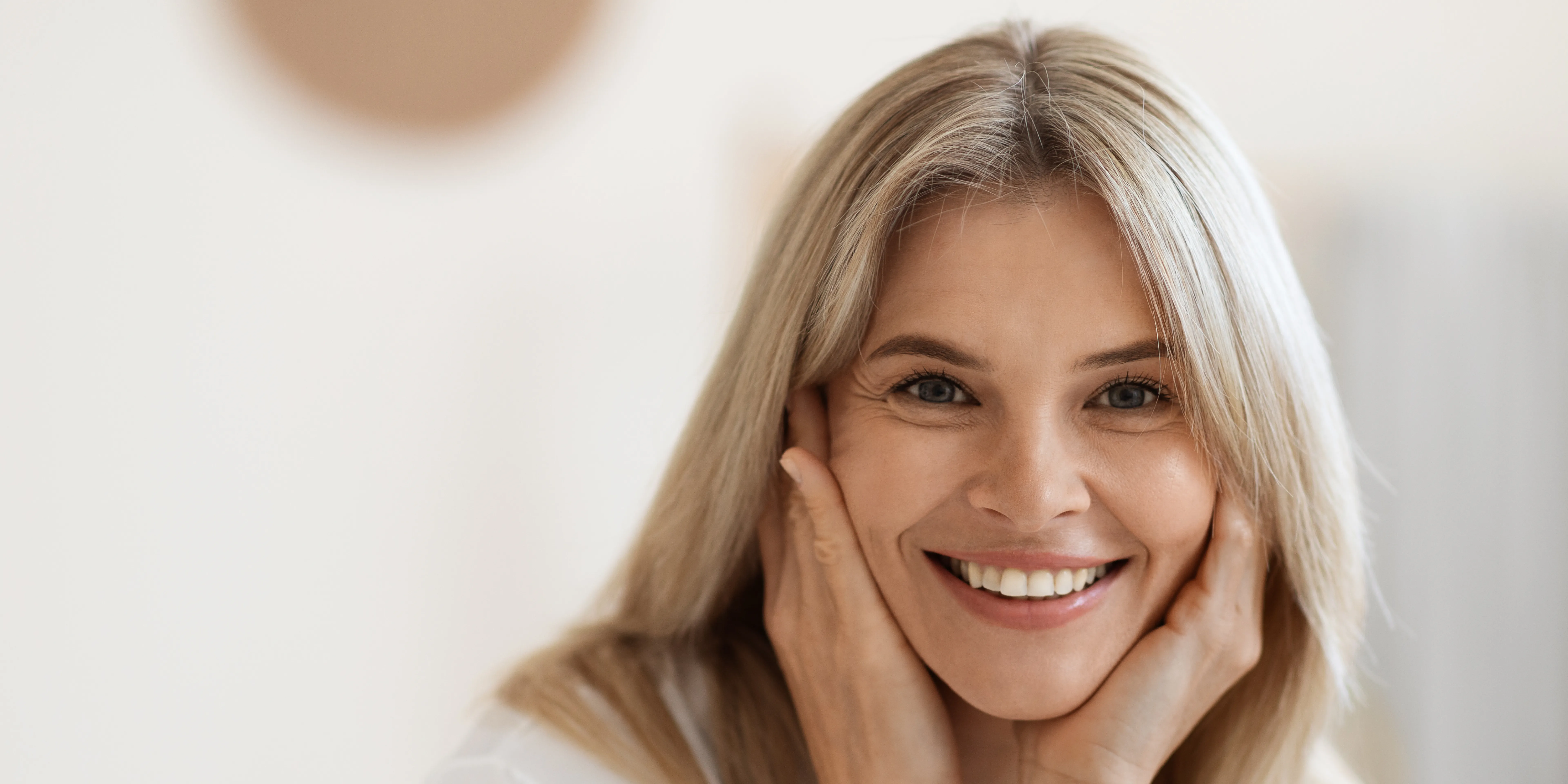 Smiling blonde woman with hands gently touching her face against a bright, neutral background.