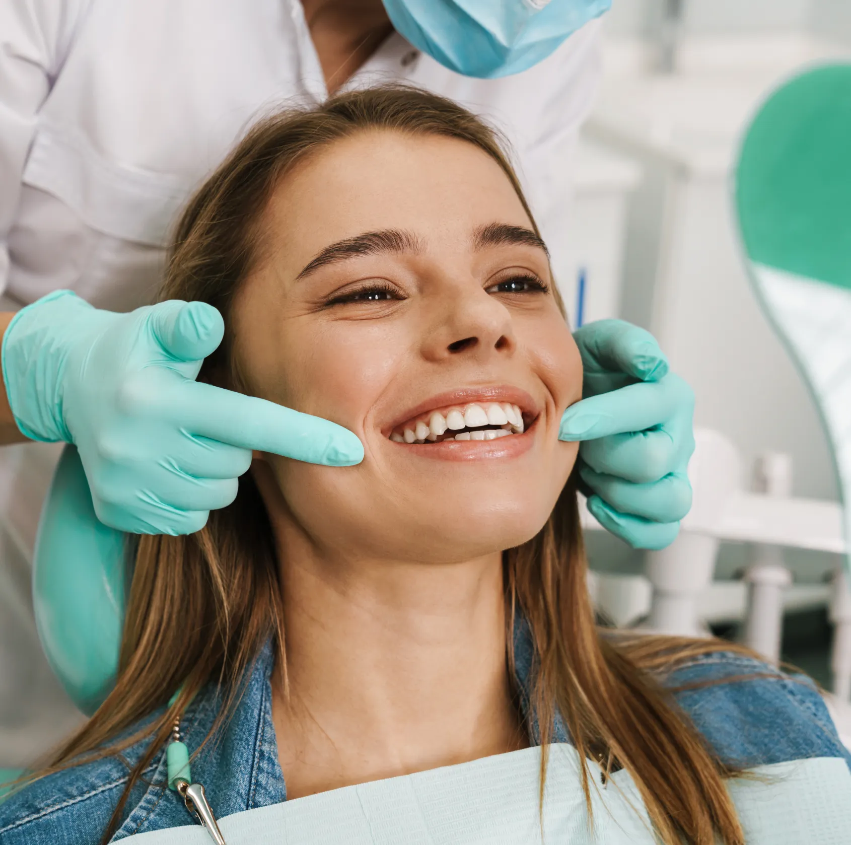 Dentist wearing gloves and mask smiling while pointing to a young woman's healthy teeth in a dental clinic.