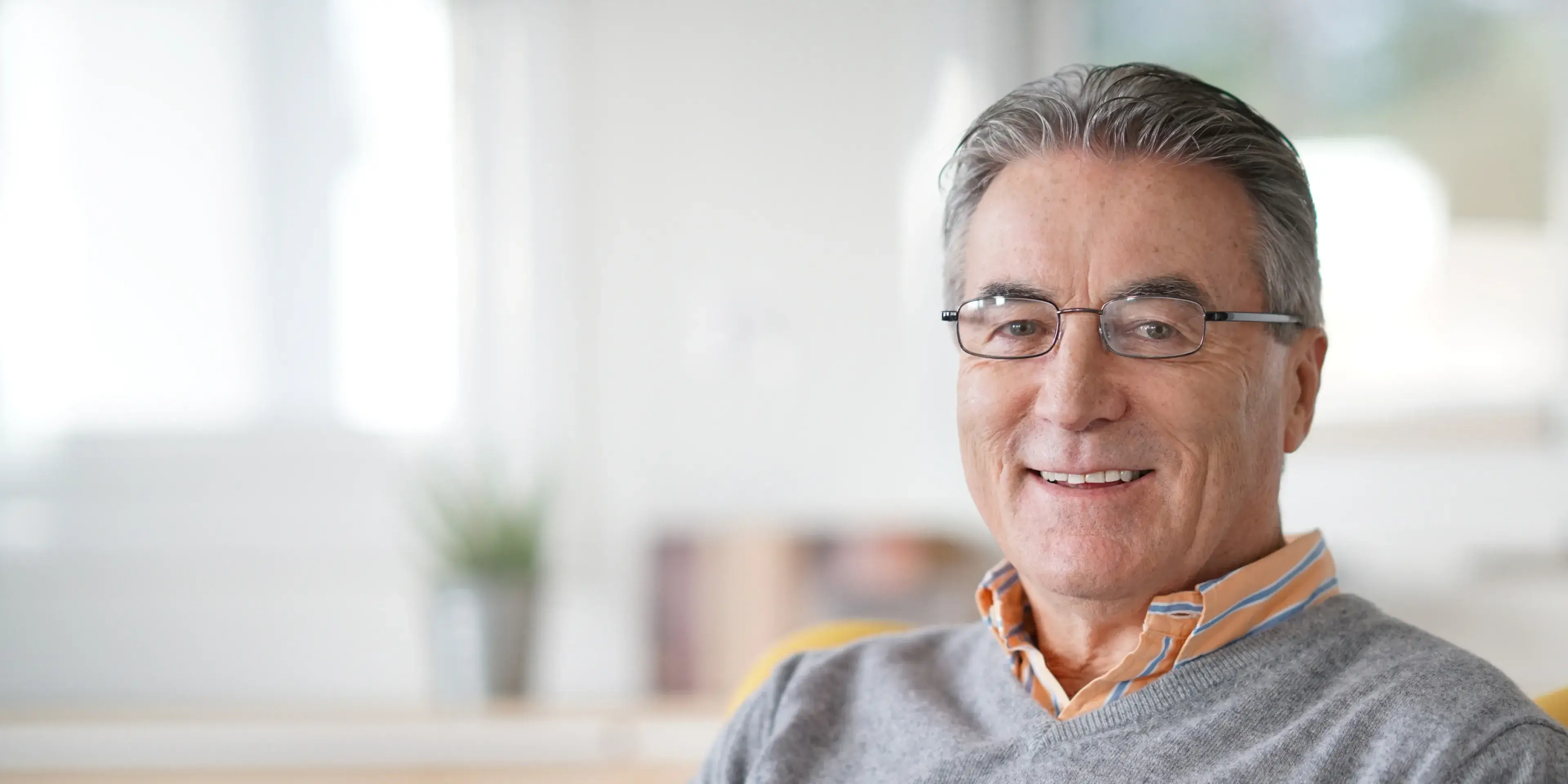 Smiling elderly man wearing glasses and a gray sweater sitting in a bright, modern room.