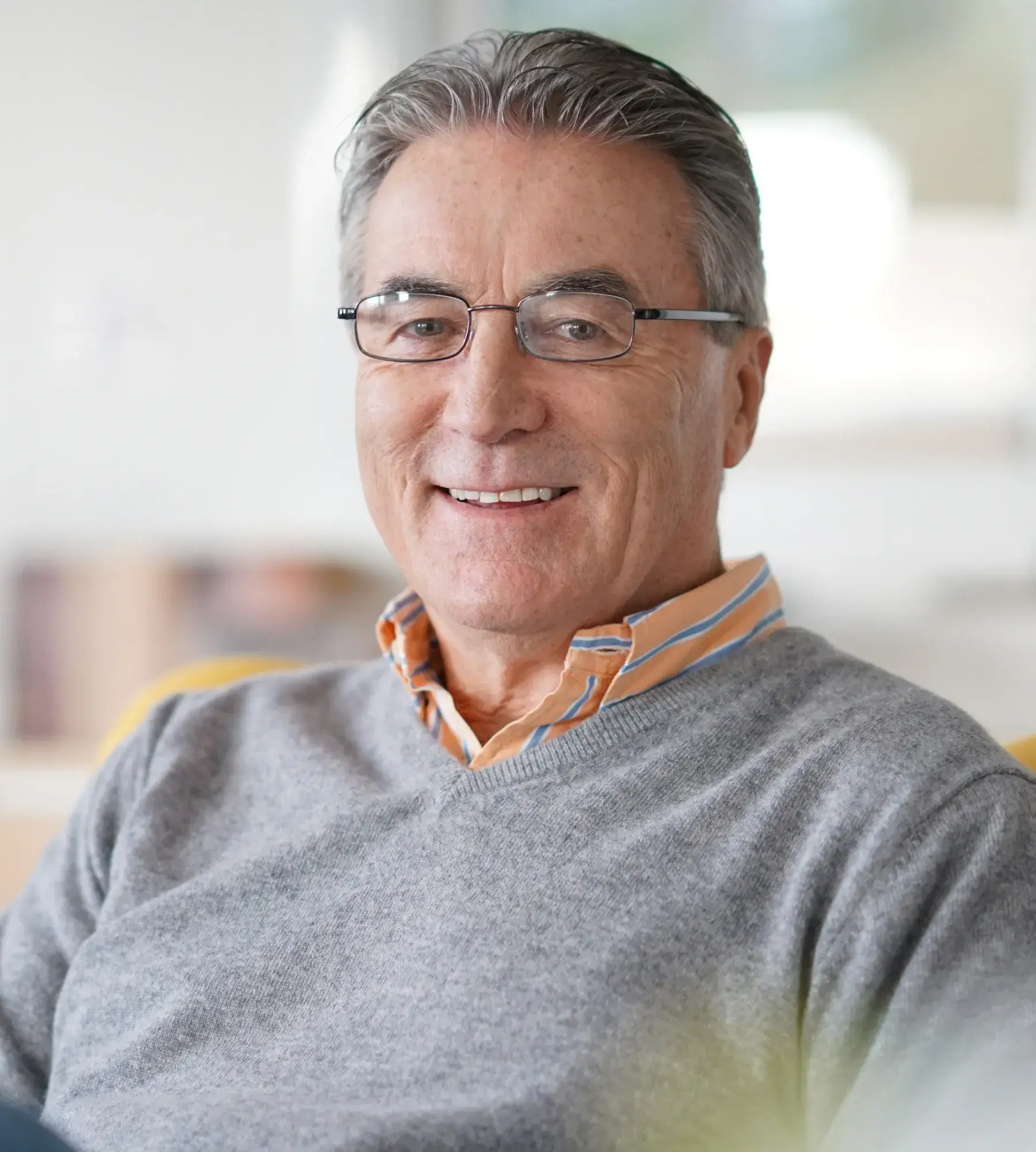 Smiling older man with glasses wearing a gray sweater over an orange striped shirt in a bright room.