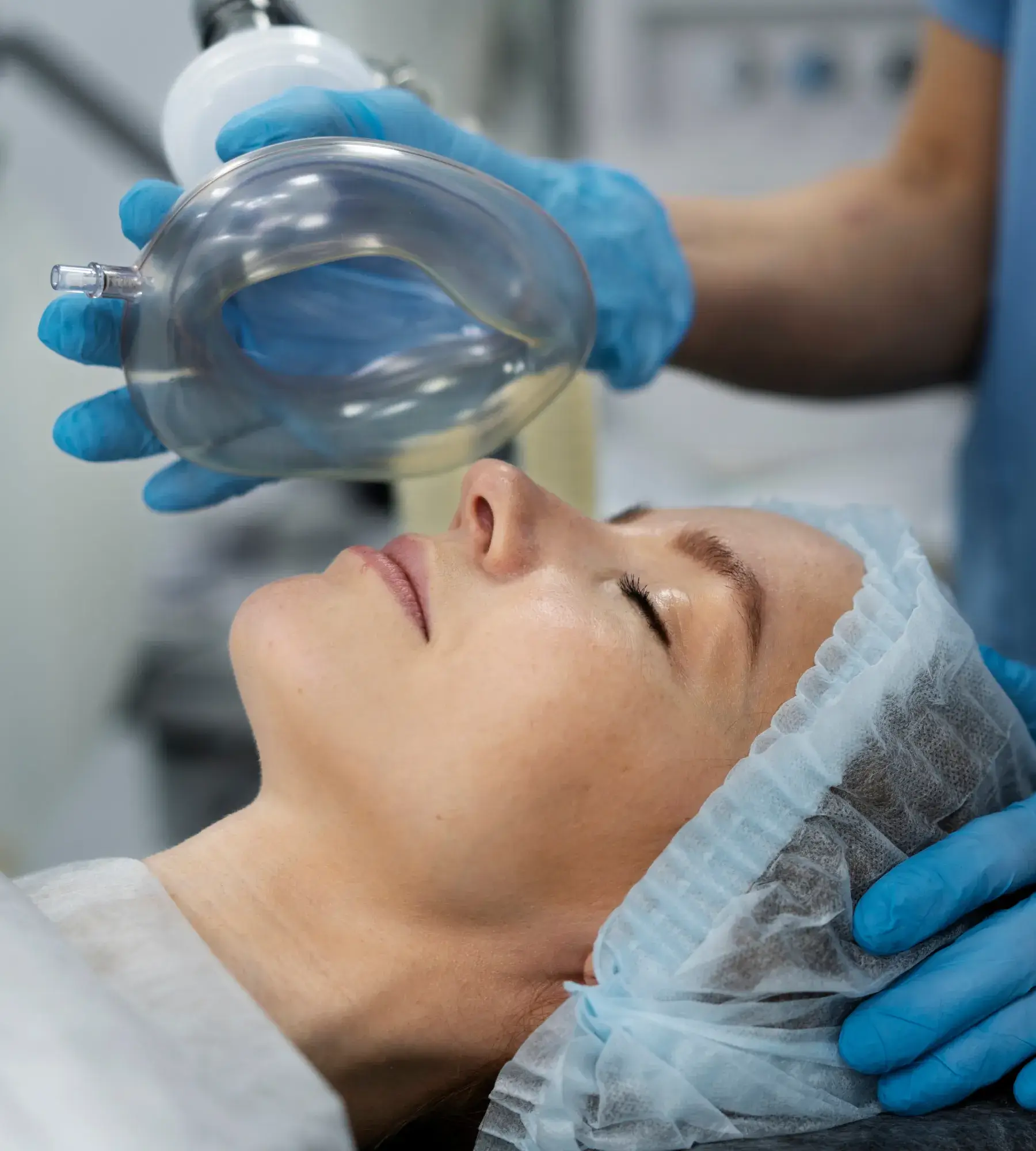 Patient wearing a surgical cap lying with eyes closed as a healthcare professional in blue gloves holds an anesthesia mask near their face.