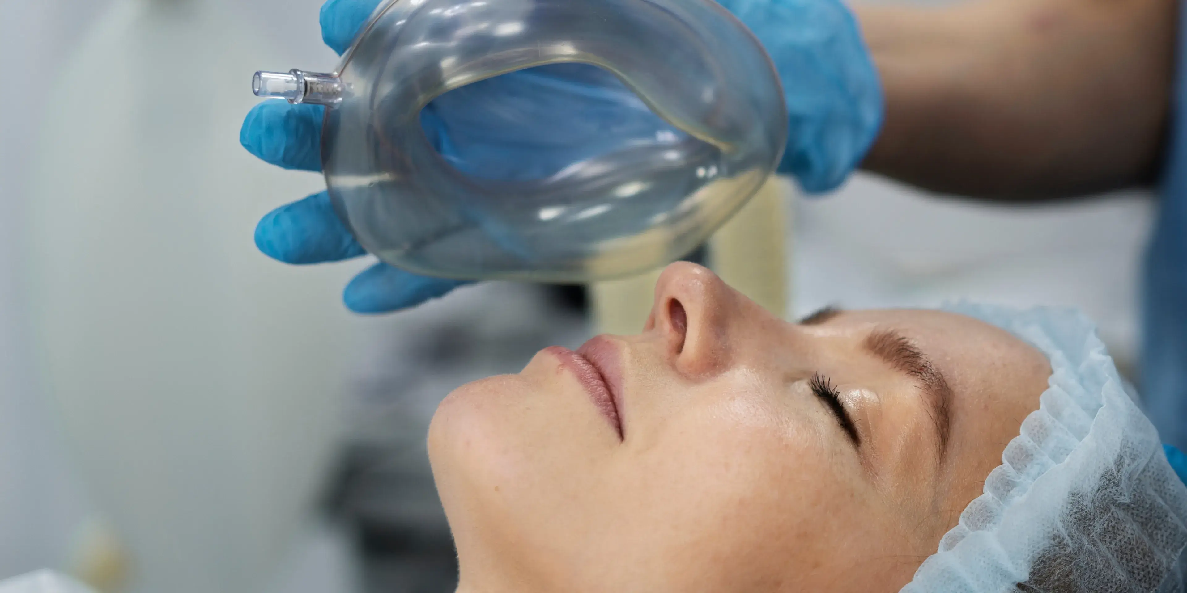 Close-up of a patient with closed eyes wearing a surgical cap while medical staff holds a clear oxygen mask near their face.