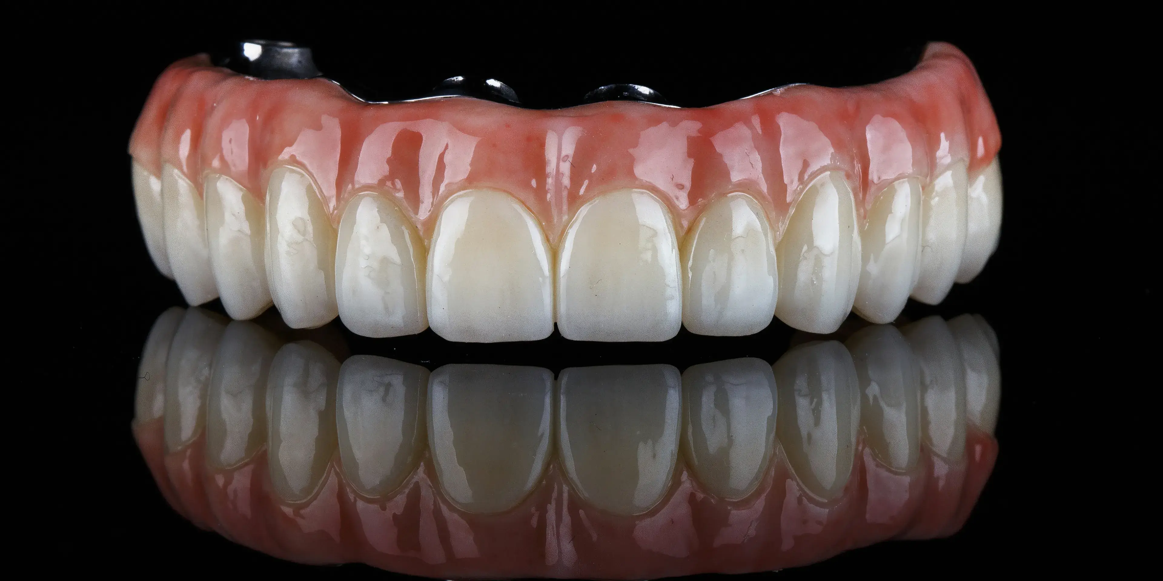 Upper dental implant bridge with natural-looking teeth and pink gum base reflected on a black surface.
