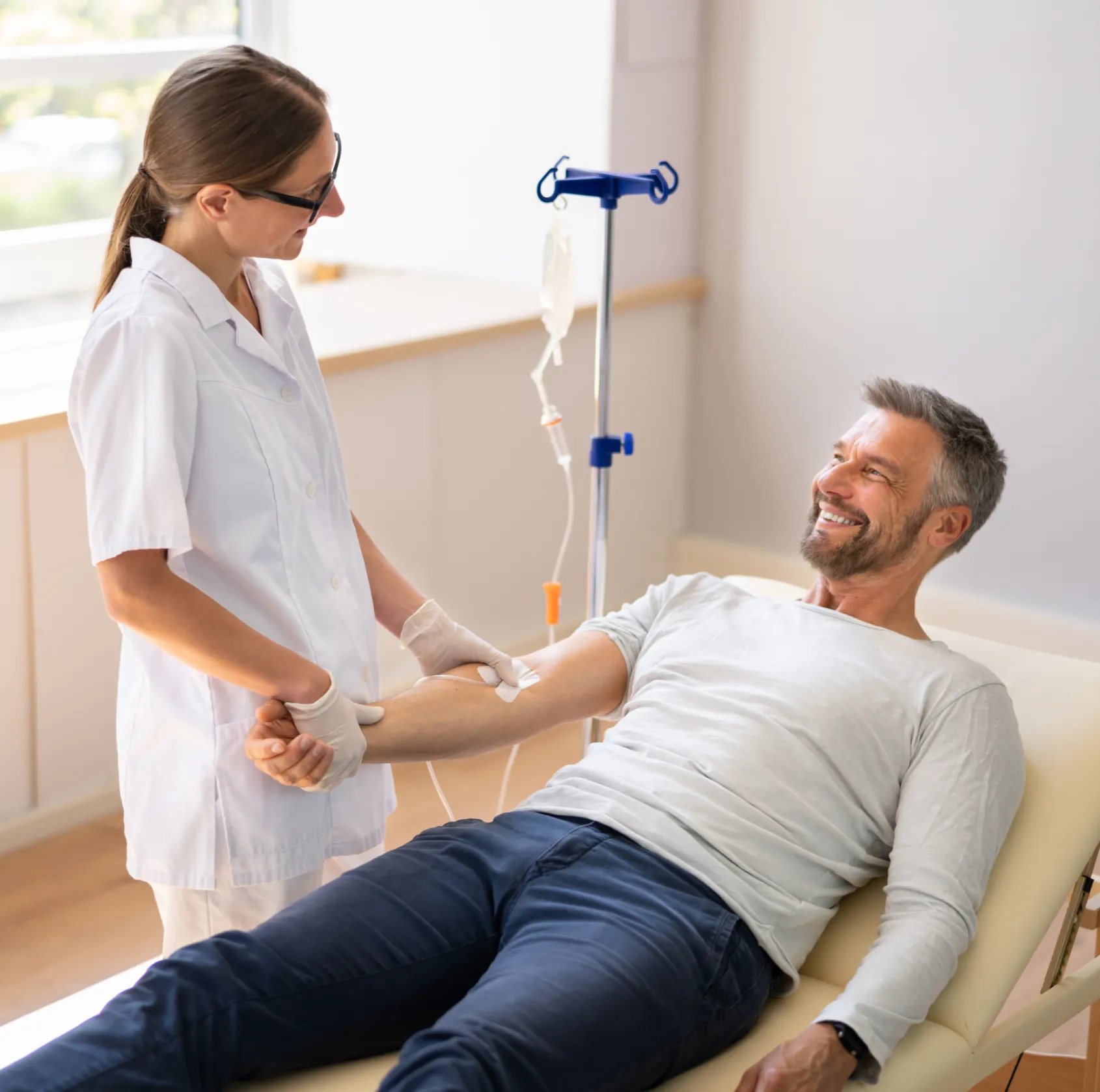 Healthcare professional in white coat and gloves attending to a smiling man receiving an intravenous drip in a clinic.