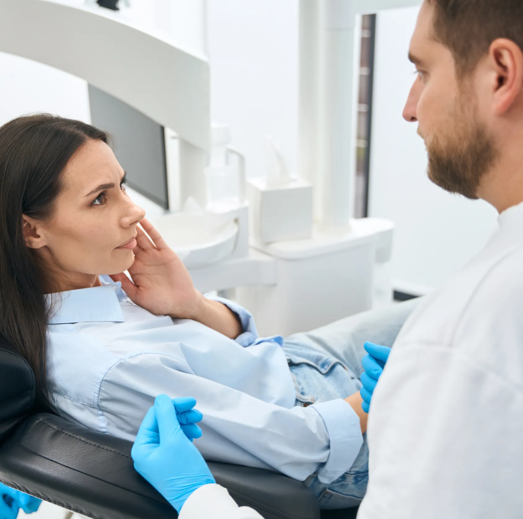 Woman sitting in dental chair holding her jaw while a male dentist wearing blue gloves talks to her.