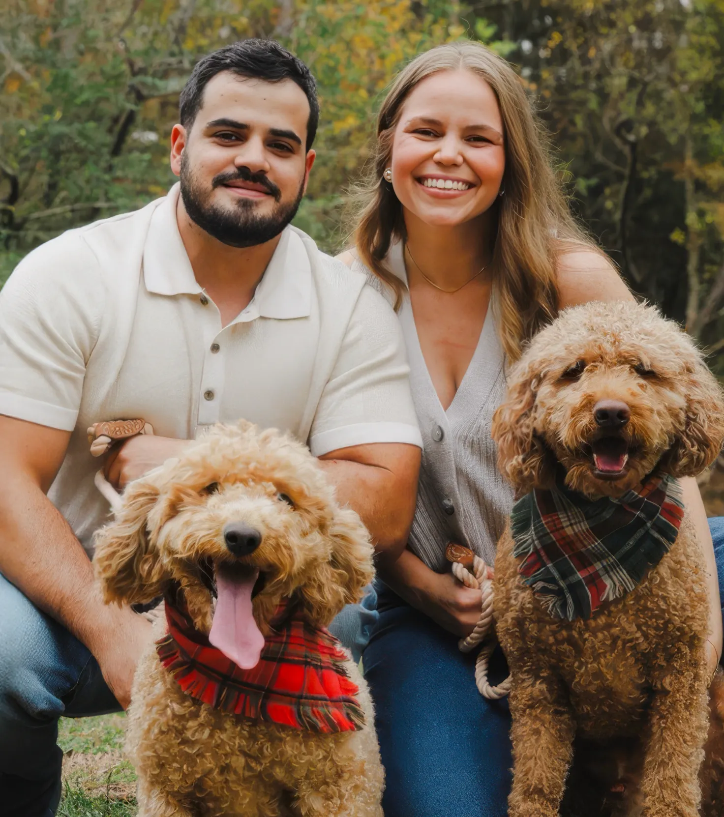 Smiling man and woman outdoors with two curly-haired dogs wearing plaid bandanas.