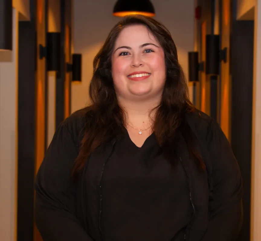 Smiling woman with long dark hair wearing a black top and necklace standing in a warmly lit hallway.