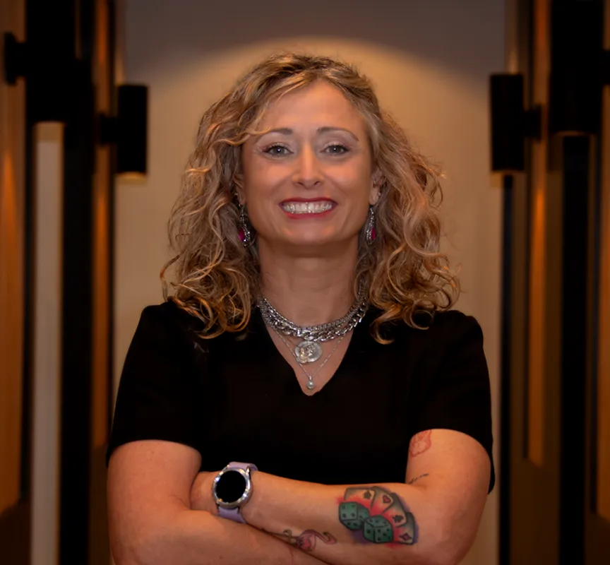 Smiling woman with curly blonde hair, wearing a black shirt, layered necklaces, earrings, a smartwatch, and dice tattoos on her arm.
