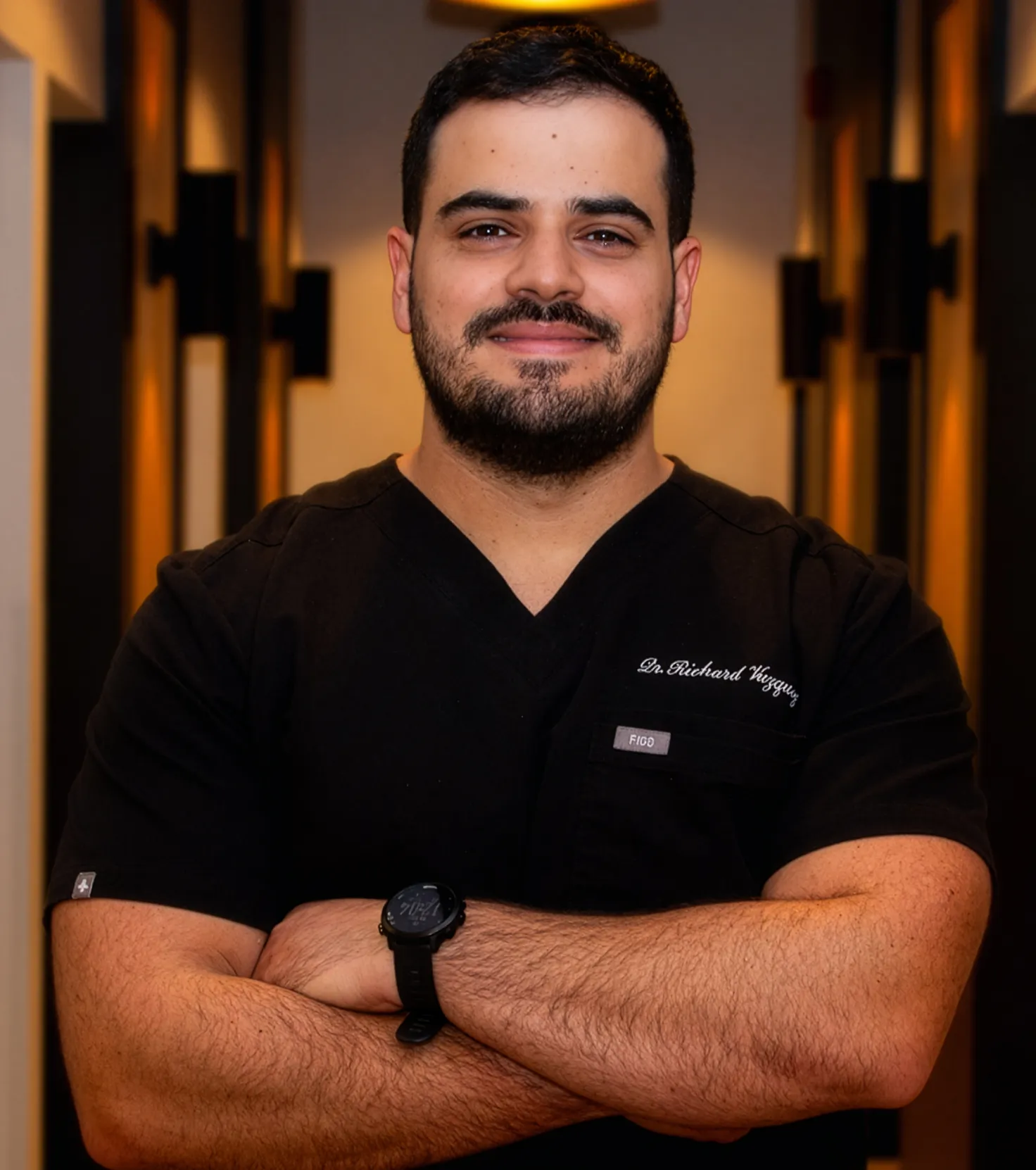 Smiling male doctor with beard wearing black scrubs and a watch, standing with arms crossed in a warmly lit corridor.