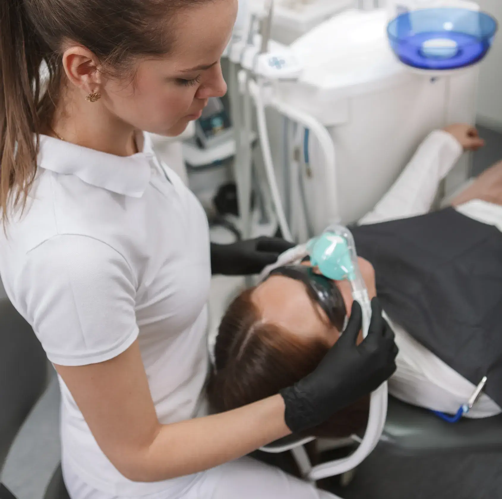 Dental professional in white uniform holding a sedation mask over a patient's face in a dental clinic.
