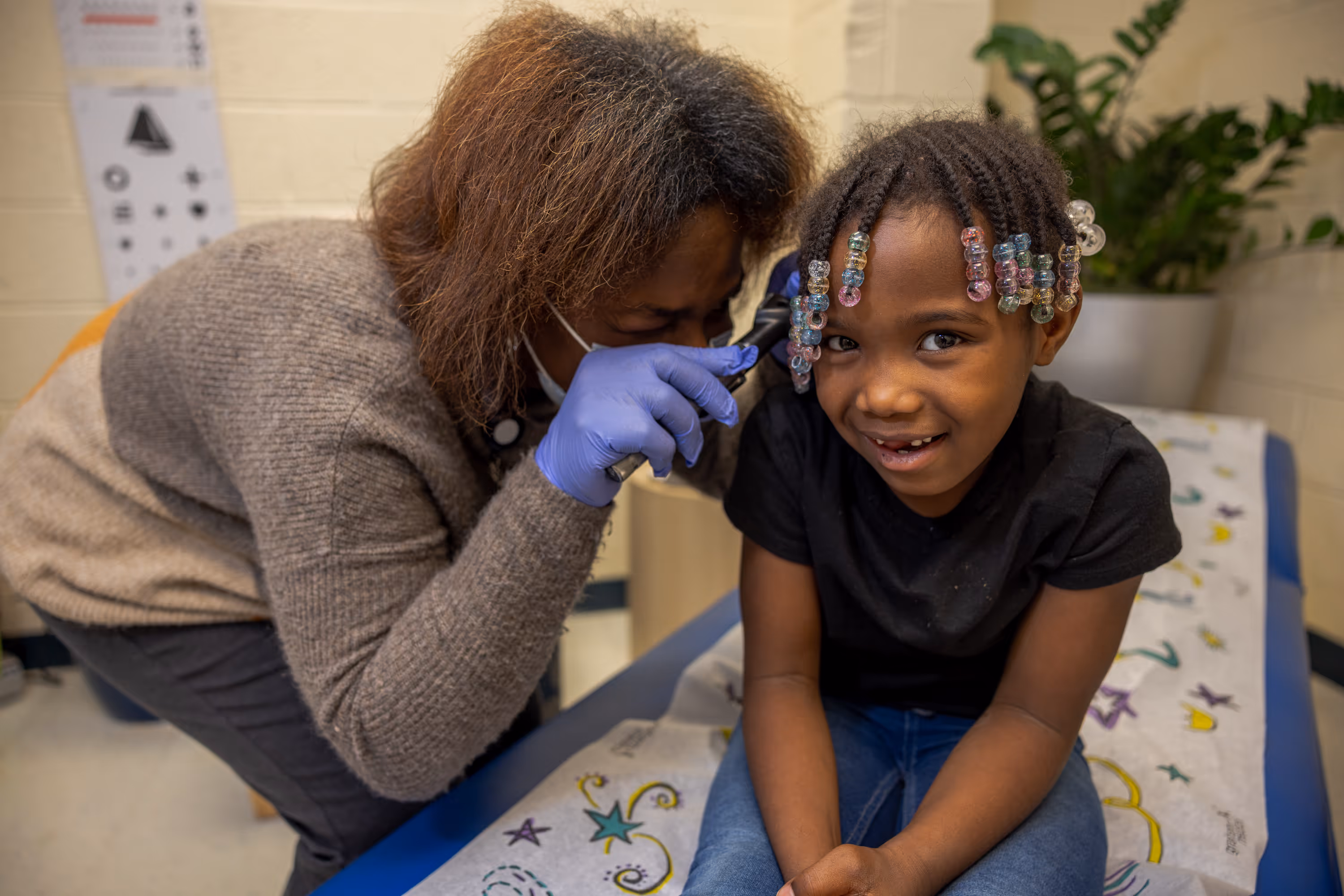 Healthcare worker wearing gloves examining ear of smiling young girl with beaded hair in clinic.
