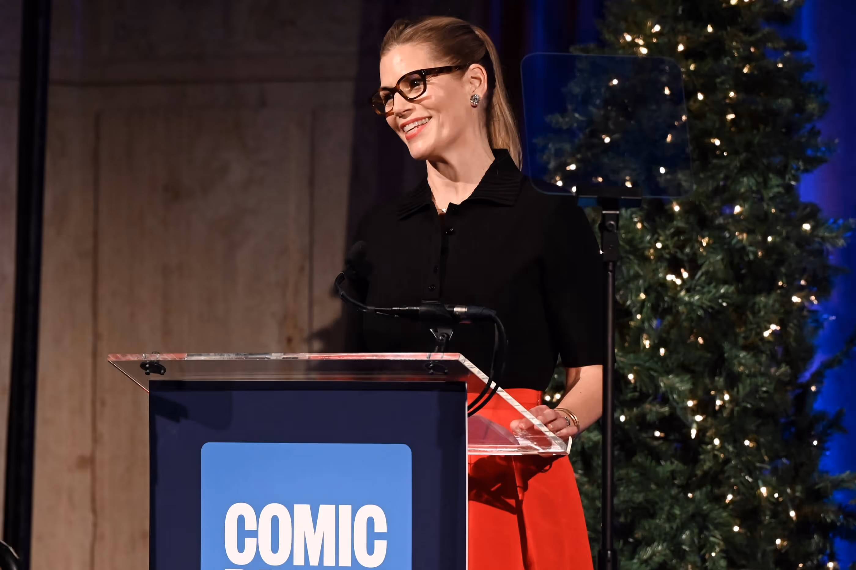 Smiling woman in black top and red skirt speaking at a Comic Relief podium with Christmas trees in the background.