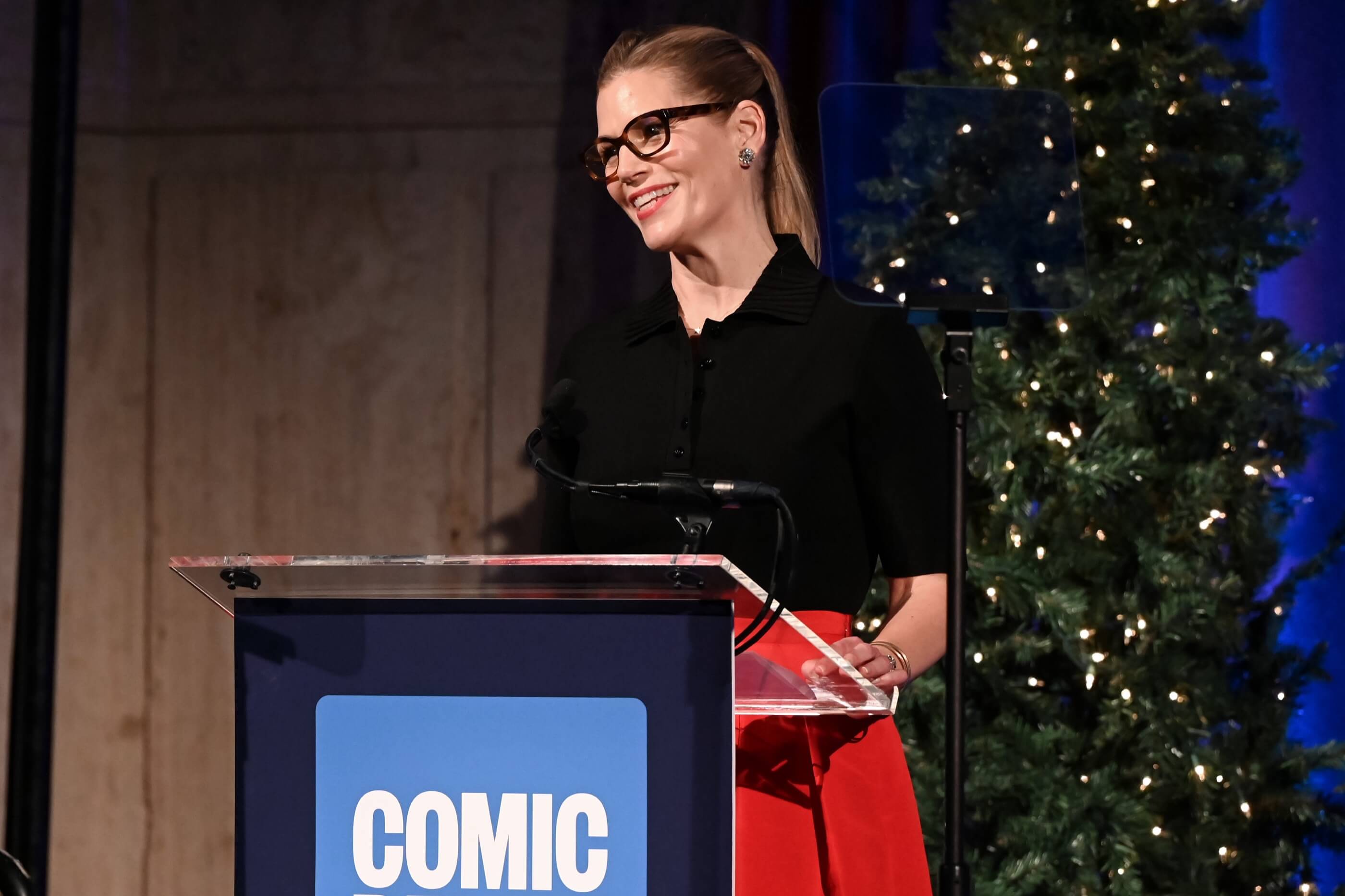 Smiling woman in black top and red skirt speaking at a Comic Relief podium with Christmas trees in the background.