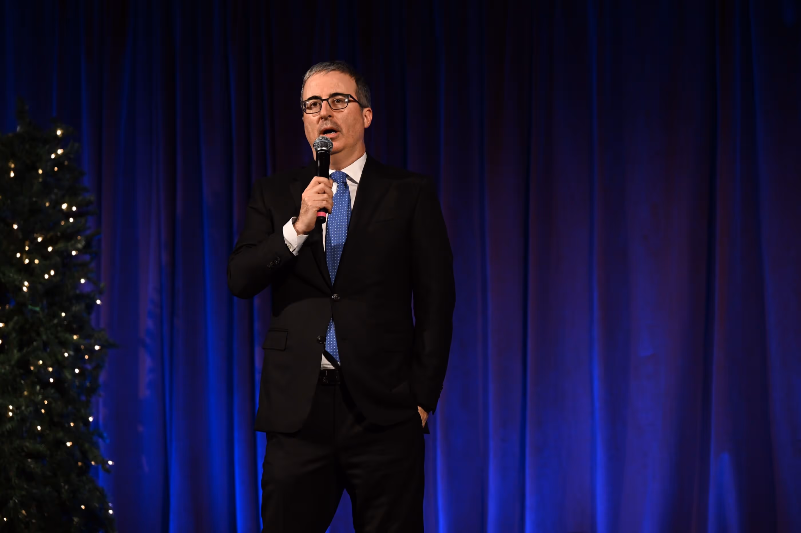Smiling young man in a suit holds a microphone while standing on a stage with blue curtains and a Christmas tree.