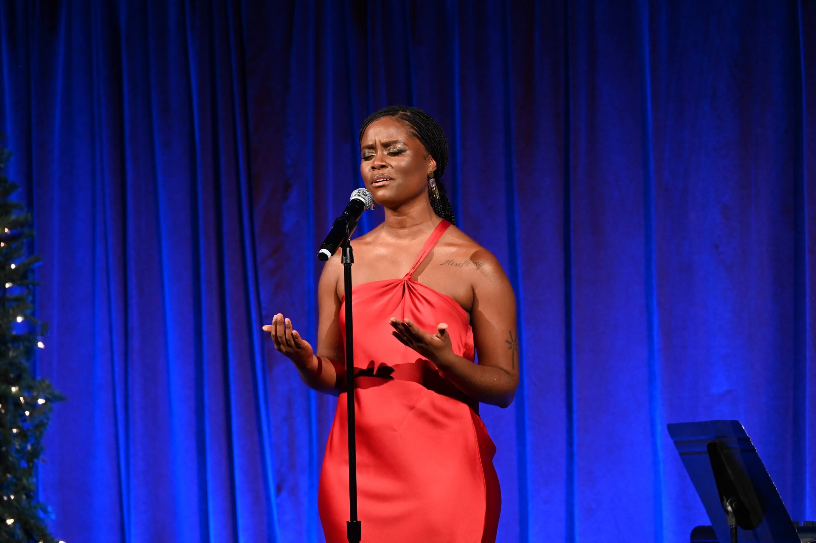 Man in glasses and a suit speaks into a microphone on a stage with blue curtains and a decorated Christmas tree.