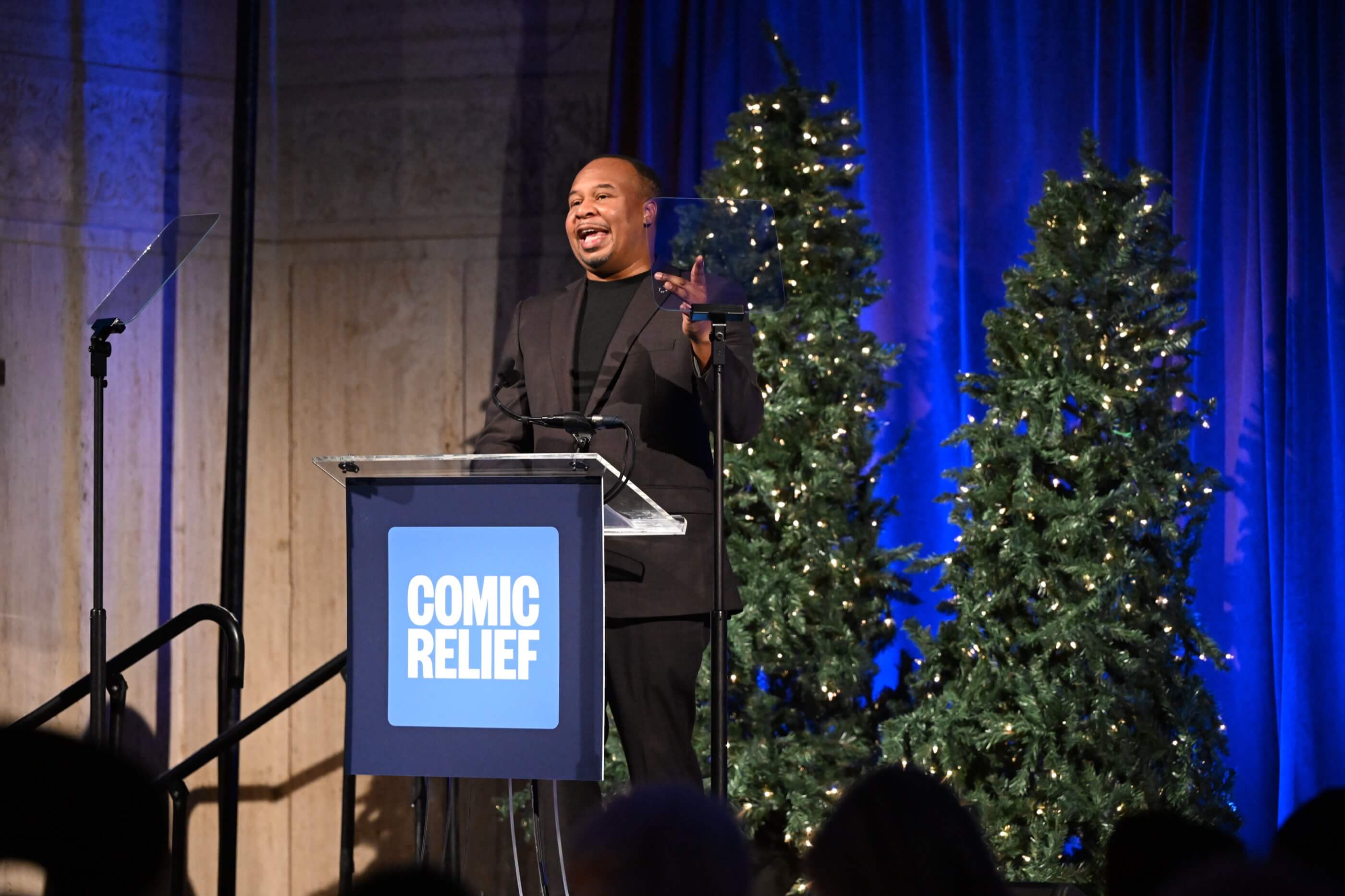 Man in black suit speaking animatedly at a Comic Relief podium with decorated Christmas trees behind him.