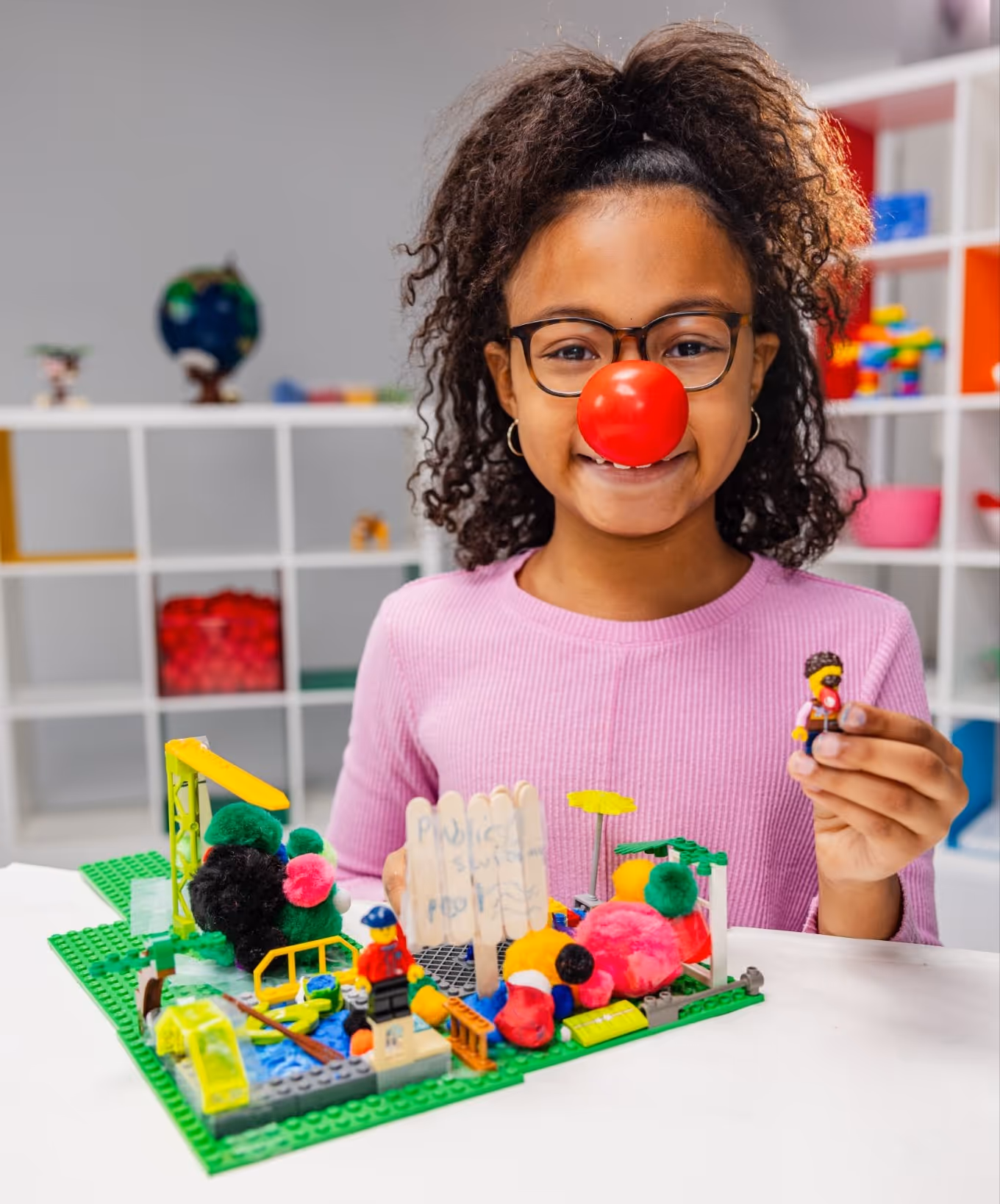 Girl with glasses and red clown nose holding a LEGO minifigure near a colorful LEGO scene with pom-poms.