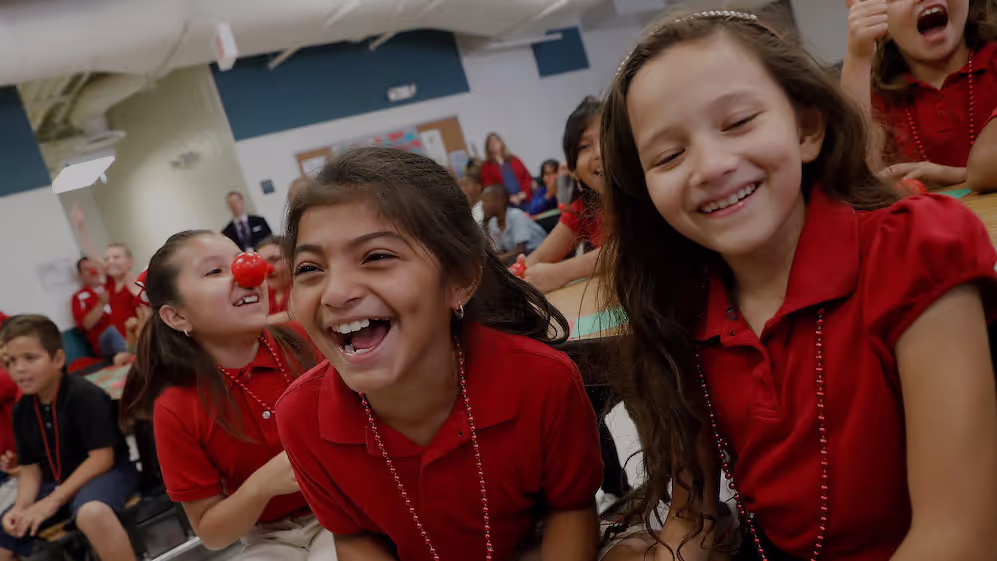 Children in red shirts with red clown noses laughing and having fun in a classroom.