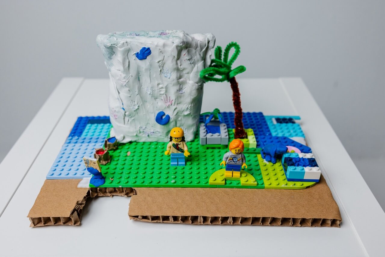 Small creative model made from Lego and craft materials including a white textured block, palm tree made of green pipe cleaners, and two Lego figurines.