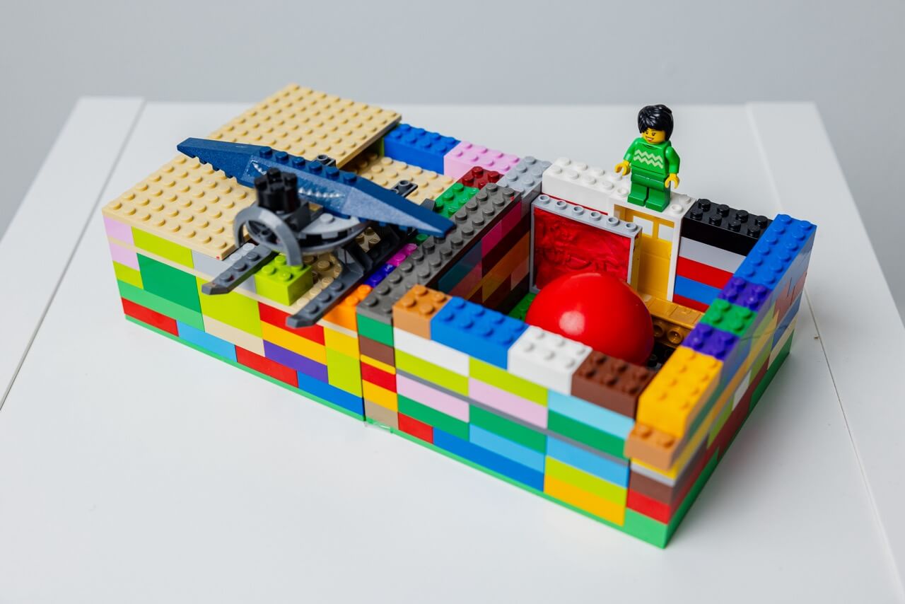 Colorful Lego structure with a large red ball inside surrounded by a block wall and a Lego figure in green standing on top.