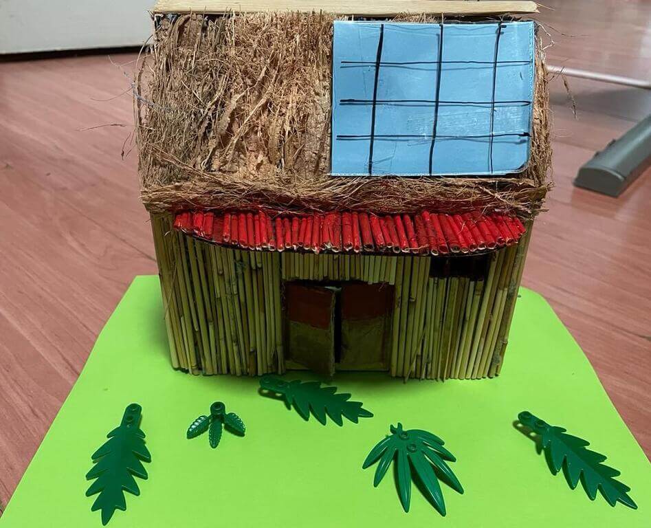 Handmade model of house with bamboo walls, a red stick roof, blue window grid, and plastic leaves on green base.