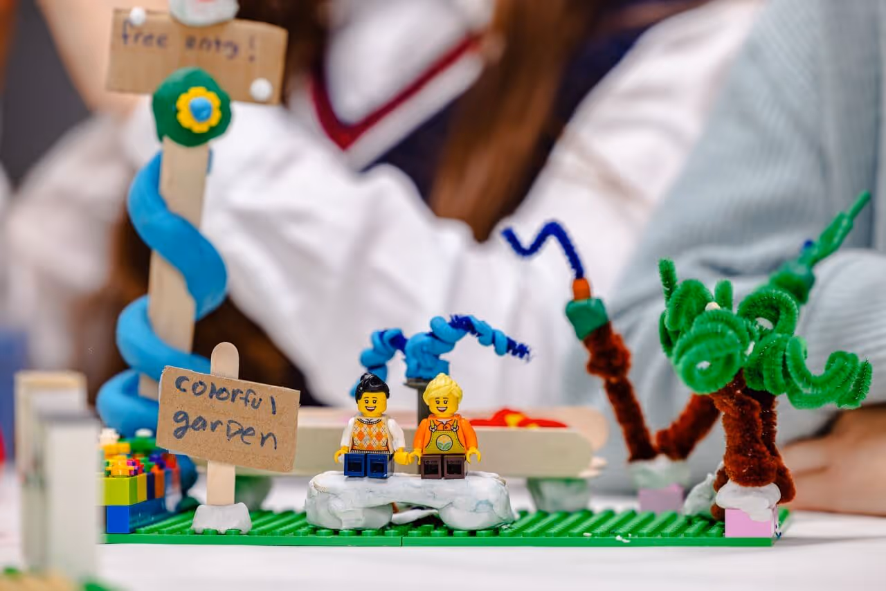 Close-up of Lego model labeled 'colorful garden' with two Lego figurines seated on white clay blocks and green pipe cleaner trees.