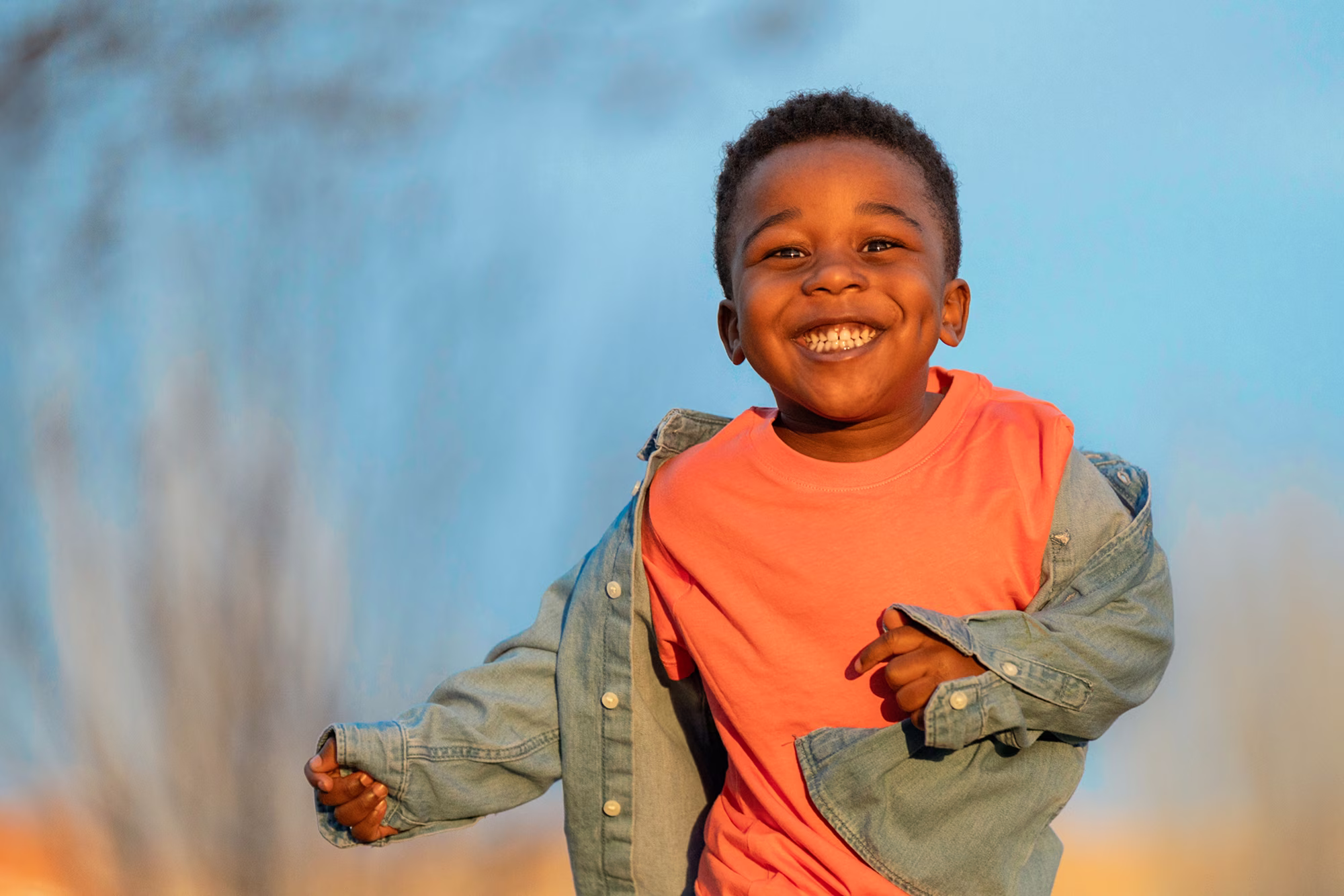 Smiling young boy wearing an orange shirt and denim jacket against a clear blue sky.