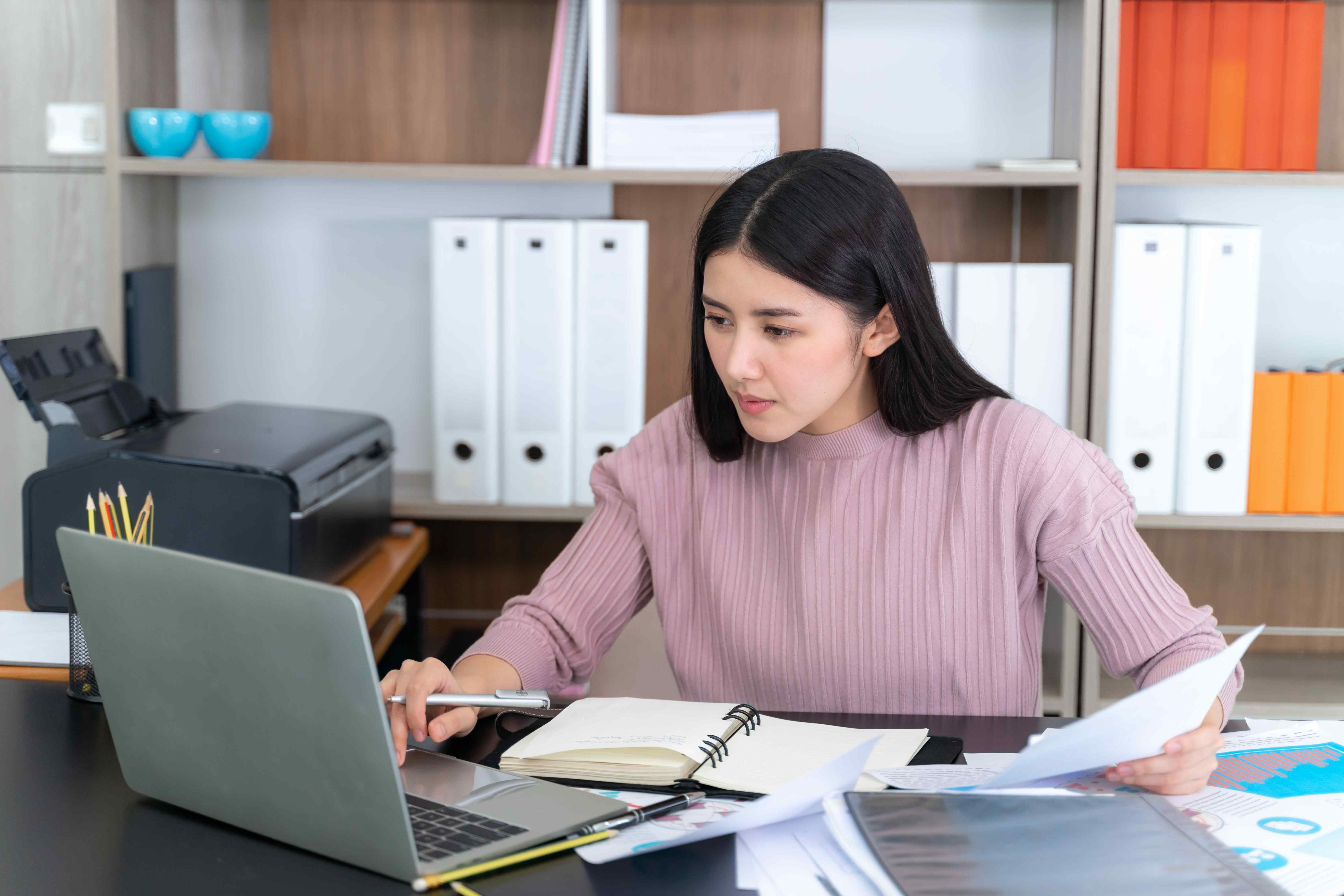 Woman working on laptop in office with documents and bookshelves
