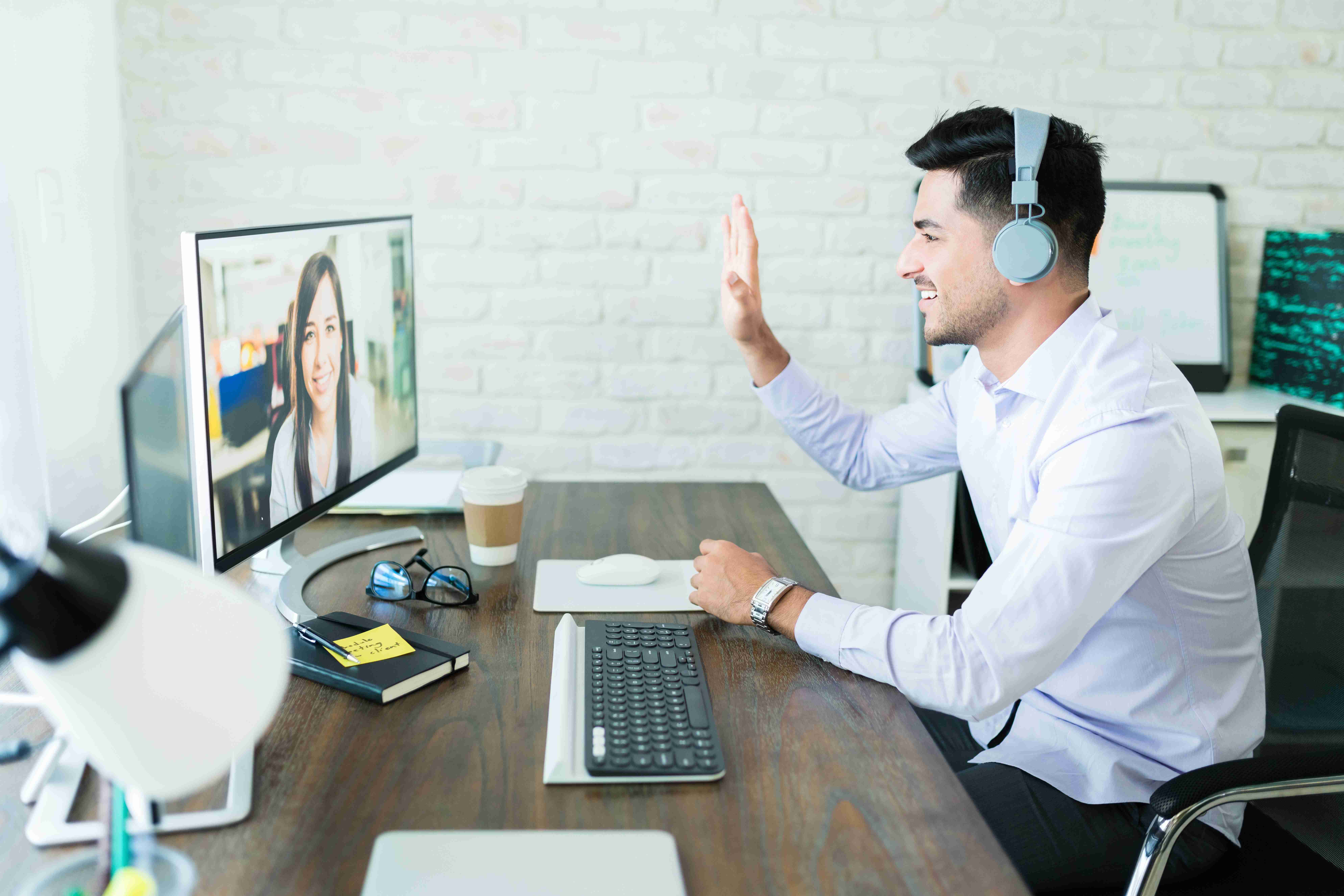 Man in headphones waves during video call at office workspace