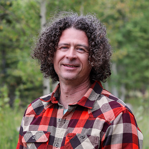 Person with shoulder‑length curly dark hair wearing a red and black plaid flannel shirt, smiling outdoors among green trees.