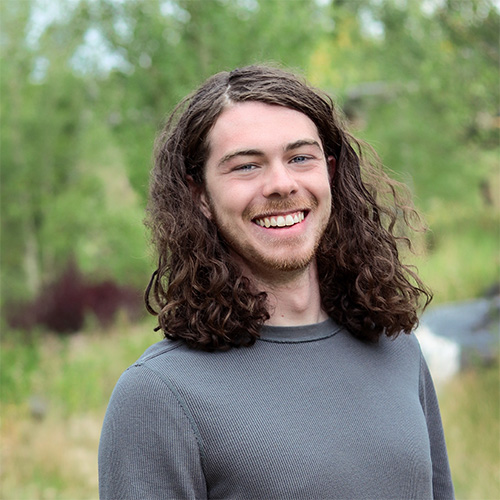 Person with shoulder‑length curly brown hair and light beard wearing a gray long‑sleeve shirt, smiling outdoors with trees and greenery in the background.
