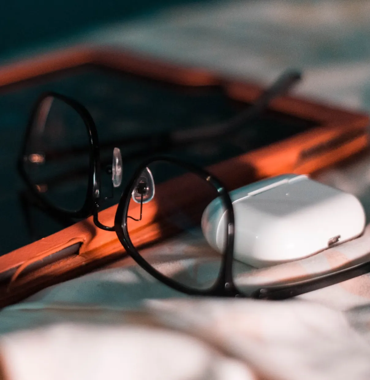 Black-framed eyeglasses resting on a white surface next to a white wireless earbud case and an orange tablet.
