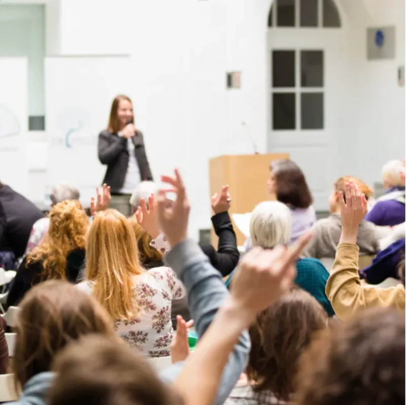 Audience with raised hands attending an in-person event with a speaker on stage.
