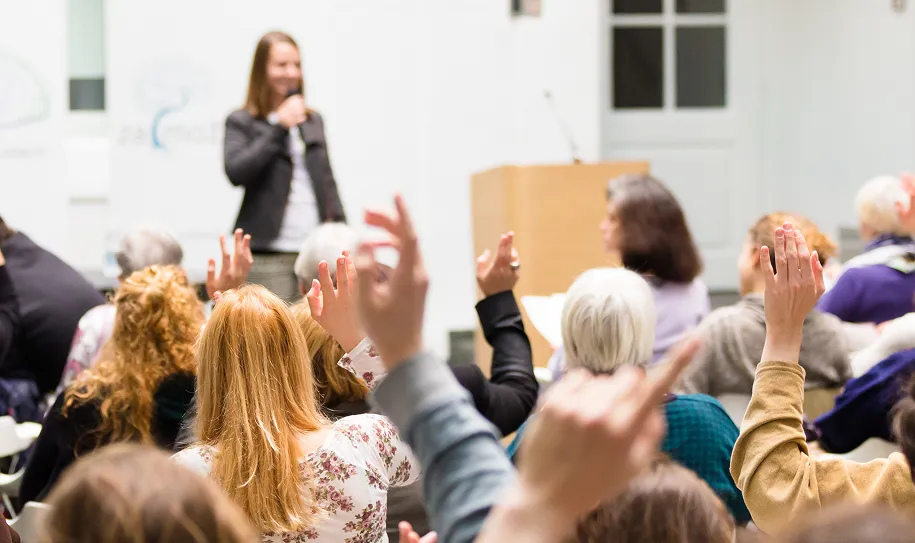 Audience members raising hands while a woman speaks on stage with a microphone.