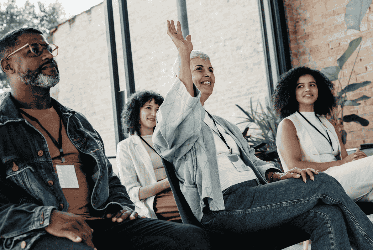 Group of diverse people seated in a casual workshop setting, with one woman raising her hand to ask a question.