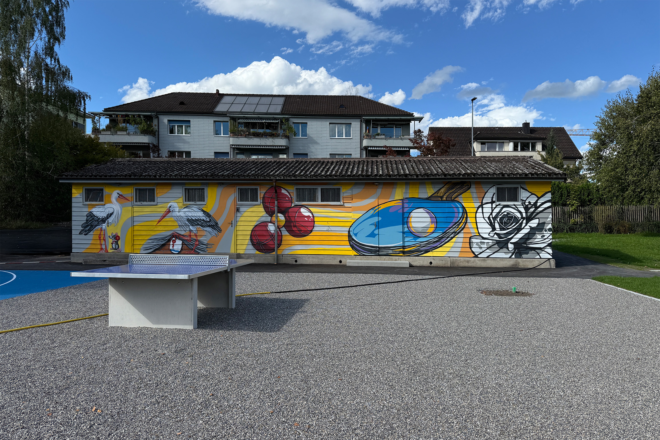Wide mural with storks, cherries, ping-pong paddle and rose set on yellow wave patterns; playful public art across the main front.