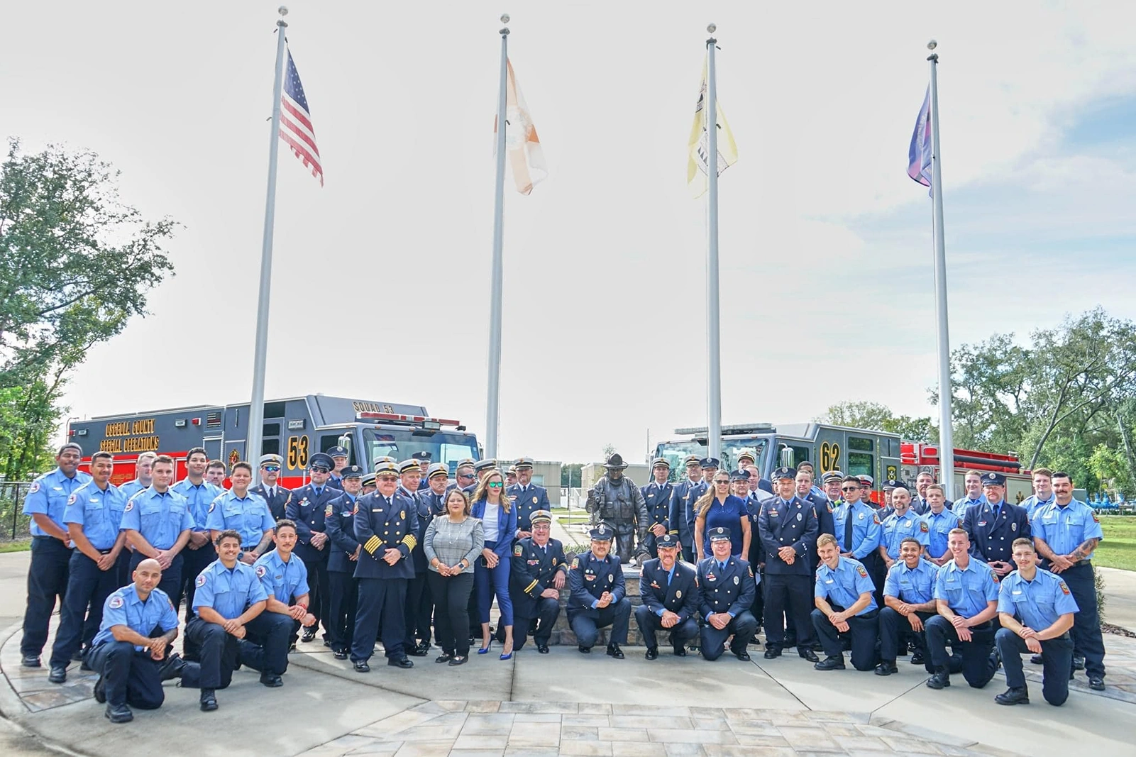 A group of Osceola firefighters and officials posing in front of fire trucks and flagpoles in a park setting. The team stands together, wearing uniforms, and there are several flags displayed in the background.