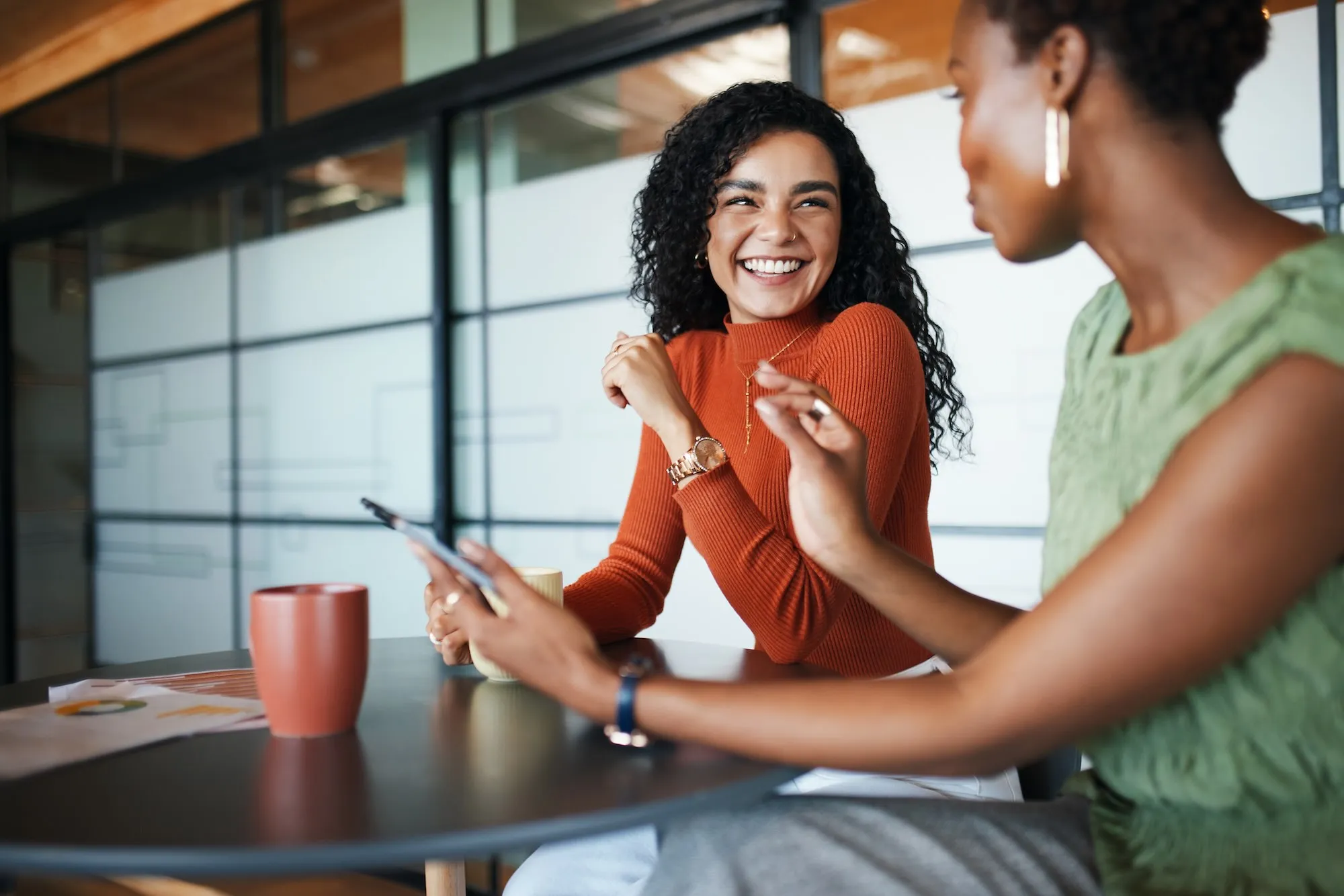 Two women having a friendly conversation over coffee at a modern office table.