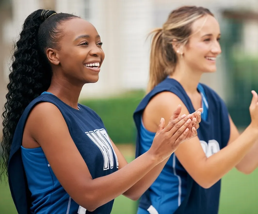 Two female athletes wearing blue sports jerseys clapping and smiling during an outdoor event.