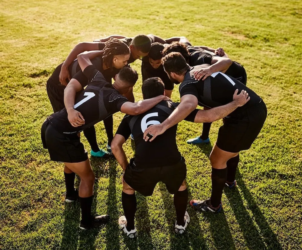 Seven male rugby players in black uniforms huddled together on green grass.