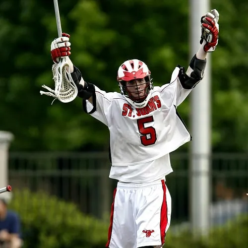Lacrosse player in white St. John's jersey raising arms with lacrosse stick in one hand during a game.