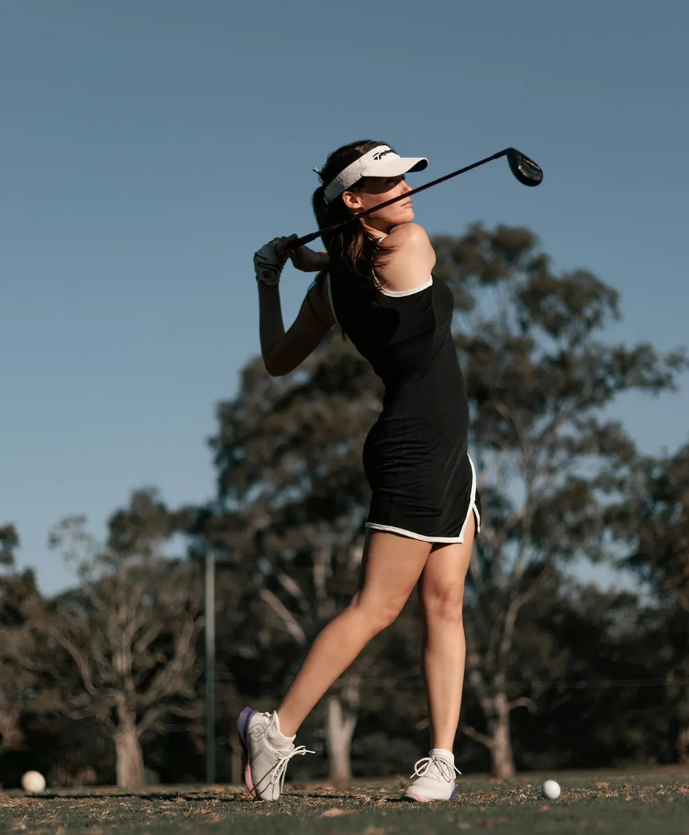 Female golfer in black dress finishing a golf swing on a course with trees in the background.